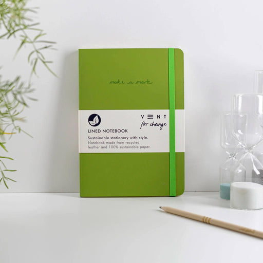 VENT sustainable A5 notebook in forest green with recycled leather cover, featuring 'make a mark' embossing and green elastic closure, shown on a white desk with minimalist styling