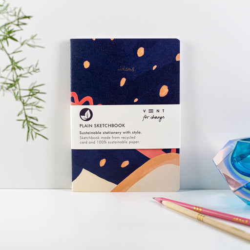 A5 sustainable sketchbook with navy blue cover featuring coral abstract patterns and dots. Product shown on white surface with decorative bamboo leaves and coloured pencils. VENT for Change branding visible on white label band.