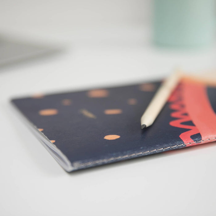 Close-up detail of VENT sketchbook's sewn binding, showing the navy blue cover with coral dots pattern and bottom edge stitching. Partial pencil visible on notebook surface.