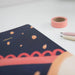 Close-up detail of navy blue sketchbook cover showing coral-coloured scalloped pattern and dots, with pink washi tape and pencils in soft focus background.