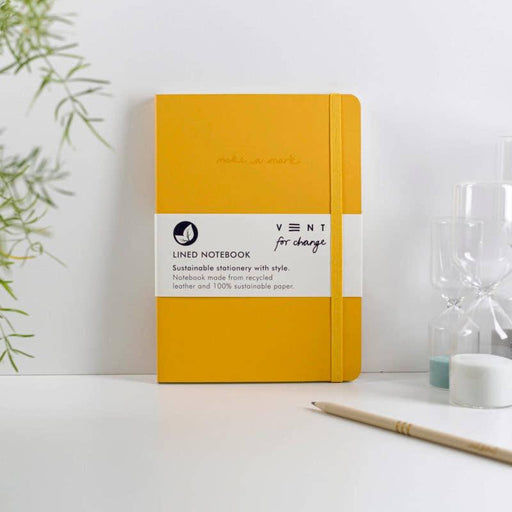 Yellow A5 recycled leather notebook by VENT on minimal white desk with bamboo plant and pencil. Sustainable stationery featuring elastic closure and "make a mark" debossed text.