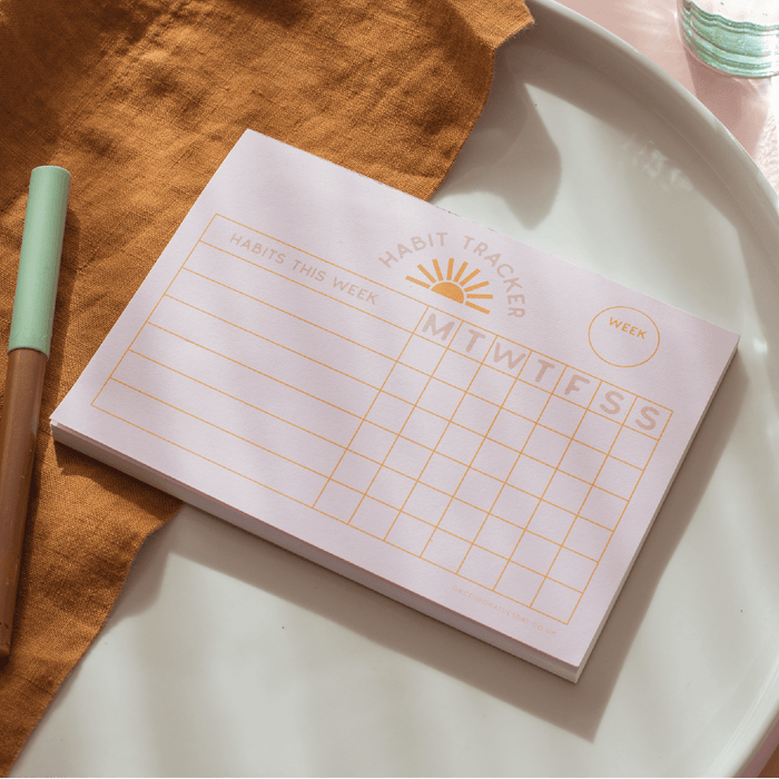 A6 Weekly Habit Tracker pad with sunrise logo and daily tracking grid, styled on white ceramic plate with mint pen and rust linen