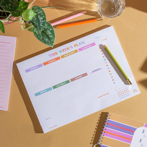 Weekly planner pad displayed on warm beige desk with colorful day tabs from Monday to Sunday. Features pastel rainbow colour scheme, habit tracker dots, and is styled with pencils, a glass of water, and a monstera plant leaf in the corner.