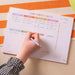 Person wearing a gingham check sleeve writing in weekly planner pad, showing active use of the daily schedule section and habit tracker. Planner features pastel colour-coded days and colourful dot tracking system.