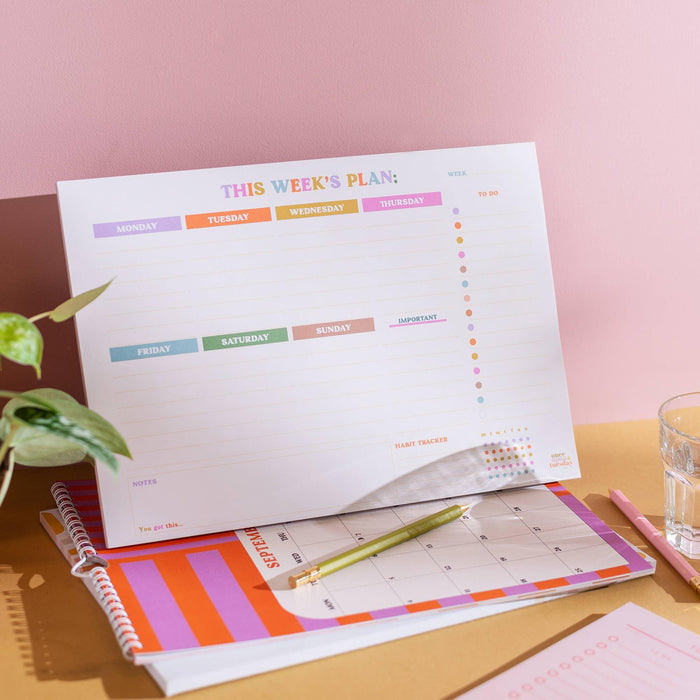 Weekly planner pad styled on desk with coordinating organisers, showing rainbow-coloured day tabs and habit tracking dots. Photographed against pink backdrop with house plant and glass of water.