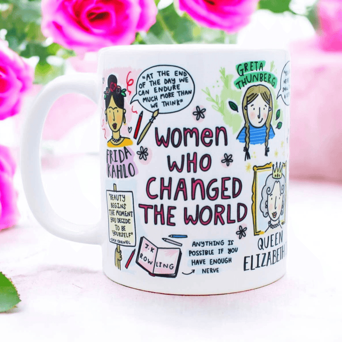 White ceramic mug featuring whimsical illustrated portraits and quotes from influential women including an artistic representation of historical figures, with pink roses in the background. The mug displays hand-drawn text reading 'Women Who Changed the World' along with inspirational quotes and decorative elements in a playful sketch style.